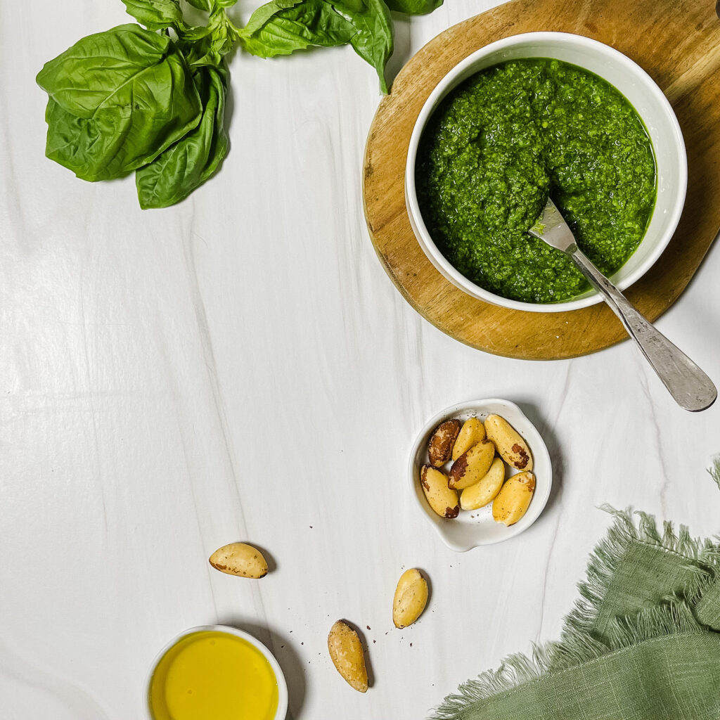 low histamine basil pesto in a white bowl, beautifully displayed on a wood platter with fresh basil leaves, and Brazil nuts and olive oil in small bowls.